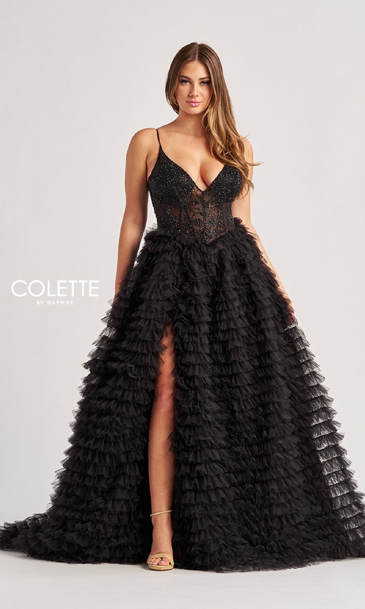 A woman wears the Long Prom Dress: Colette CL8330, a black sleeveless gown with a tiered ruffled skirt, sheer embroidered bodice, and thigh-high slit, standing against a plain light background.